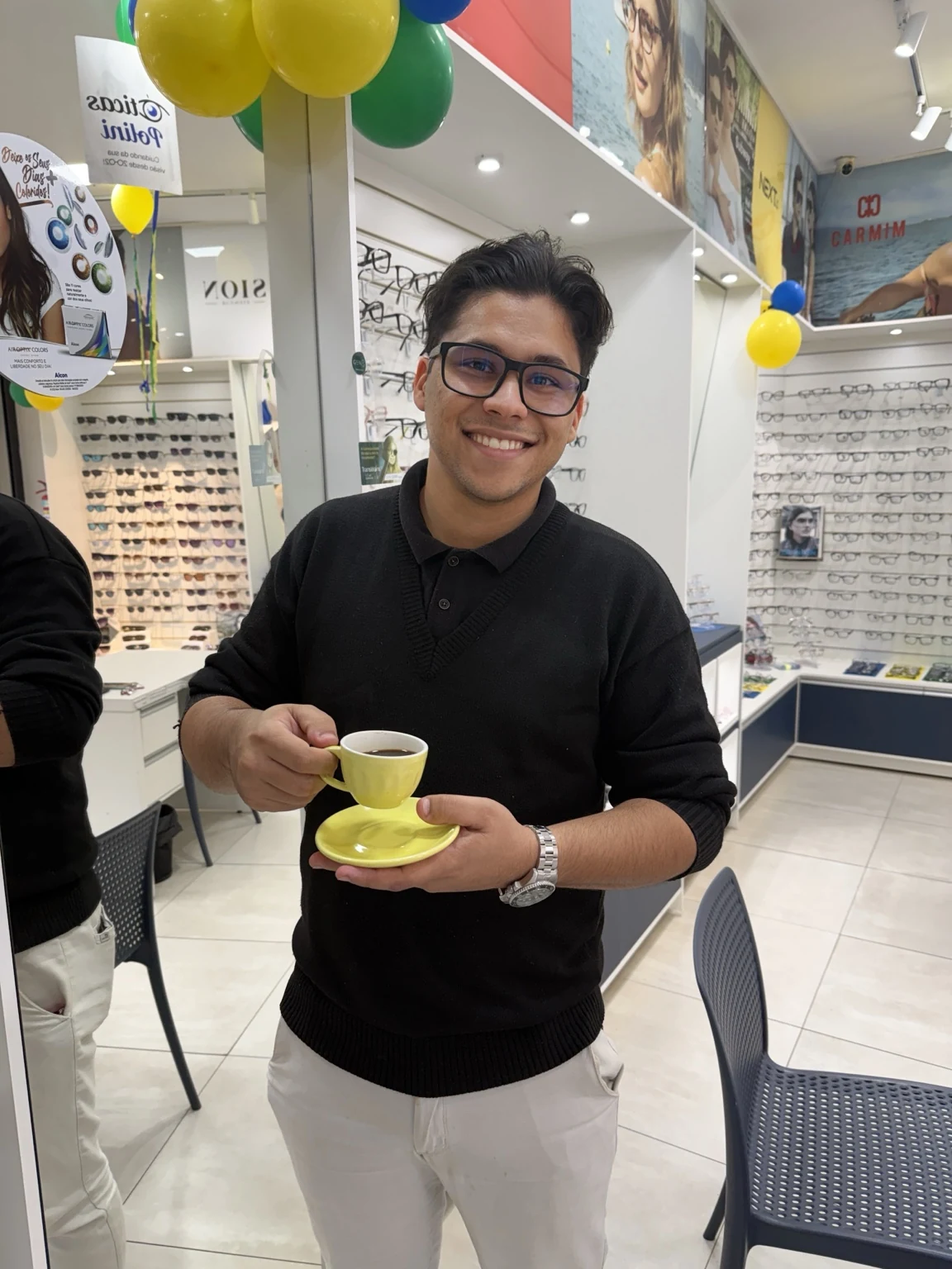 Augusto em uma ótica, sorrindo e segurando uma xícara de café durante visita profissional