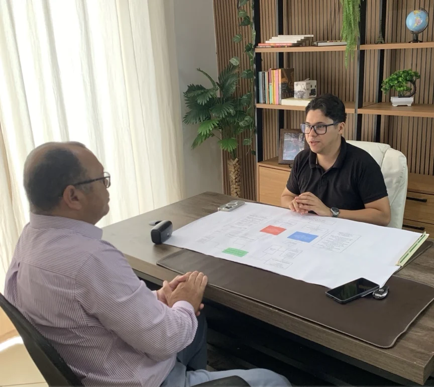 Augusto em reunião presencial de estratégia, conversando com um empresário ao redor de uma mesa com planejamento impresso